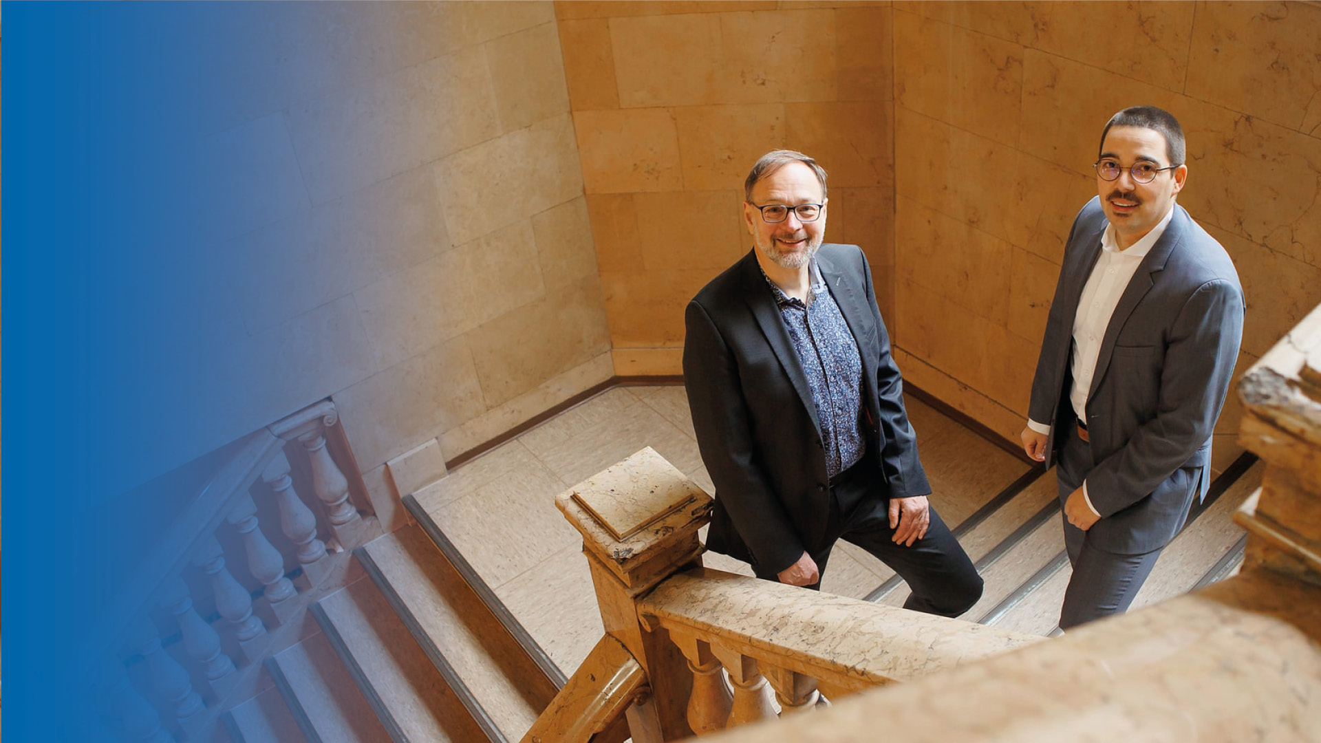 Die Arbeitsrechtsanwälte Herrn Kröll und Herr Weber in Anzügen auf einer Treppe, nach oben zur Kamera lächelnd.